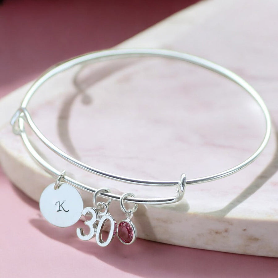 Image shows milestone birthday bangle with initial charm with the letter 'k', '30' number charm and October Rose Swarovski crystal birthstone charm.