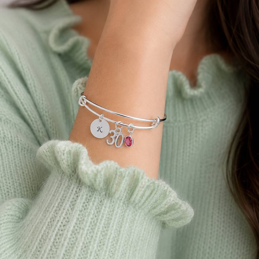 Silver charm bangle styled with an initial disc, 30 charm and pink birthstone crystal on the wrist
