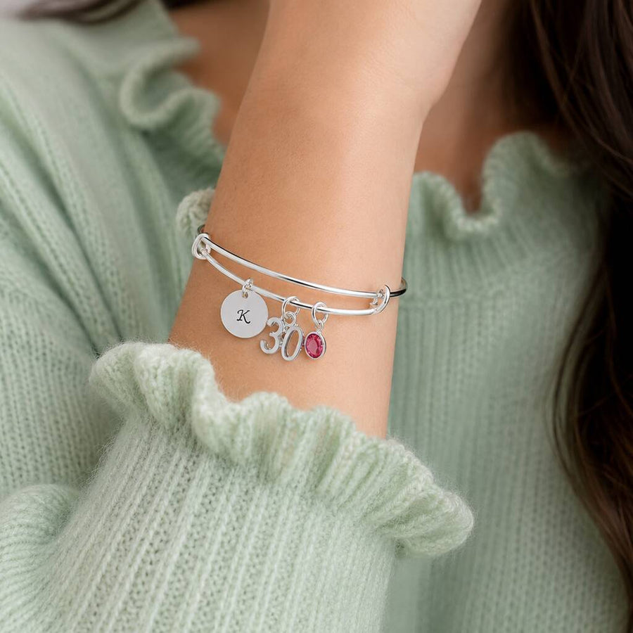 Silver charm bangle styled with an initial disc, 30 charm and pink birthstone crystal on the wrist