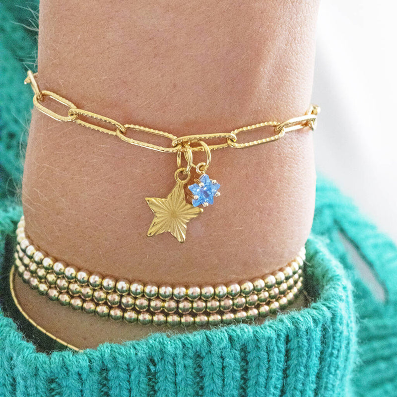 Image shows model in turquoise cardigan wearing a gold plated star charm bracelet with star birthstone detail.