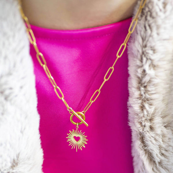 A gold sunburst charm toggle necklace worn on a pink top and fur jacket