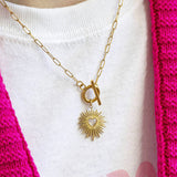 A gold sunburst charm suspended from a gold toggle necklace on a paperclip style chain worn  on a white t-shirt and bright pink cardigan