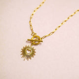 A gold sunburst charm suspended from a gold toggle necklace on a paperclip style chain laid flat on a pink back drop