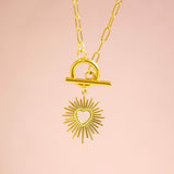 A gold sunburst charm suspended from a gold toggle necklace on a paperclip style chain