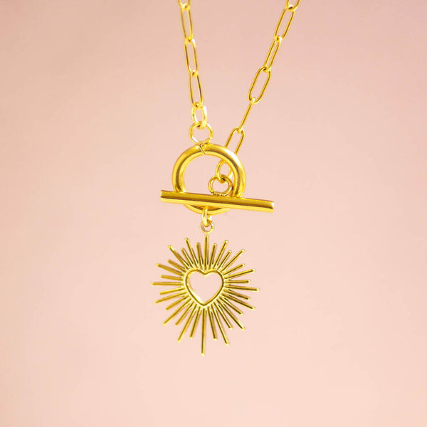 A gold sunburst charm suspended from a gold toggle necklace on a paperclip style chain