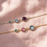 Two delicate chain bracelets with round gemstone settings, displayed on a soft pink fabric background. One bracelet is gold-toned with a green stone, a light blue stone, and a dark purple stone.