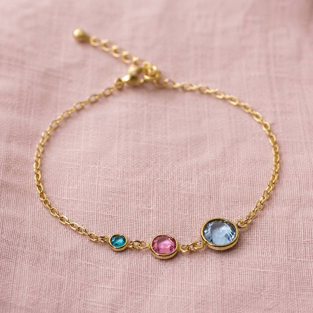 A delicate gold chain bracelet with three small bezel-set gemstones in turquoise, pink, and light blue, displayed on a textured pink fabric background.
