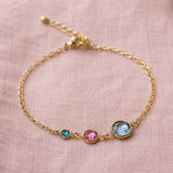 A delicate gold chain bracelet with three small bezel-set gemstones in turquoise, pink, and light blue, displayed on a textured pink fabric background.