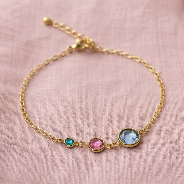 A delicate gold chain bracelet with three small bezel-set gemstones in turquoise, pink, and light blue, displayed on a textured pink fabric background.