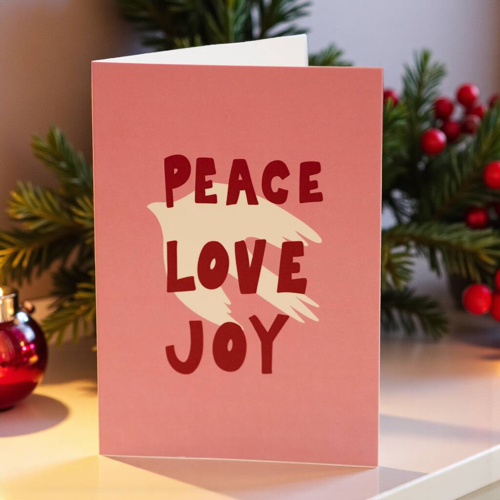 Pink greeting card with a cream dove silhouette and bold red text reading ‘Peace Love Joy’ on the front.