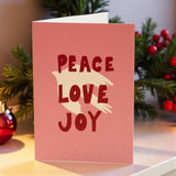 Pink greeting card with a cream dove silhouette and bold red text reading ‘Peace Love Joy’ on the front.