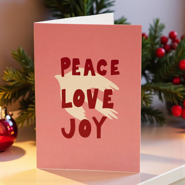Pink greeting card with a cream dove silhouette and bold red text reading ‘Peace Love Joy’ on the front.