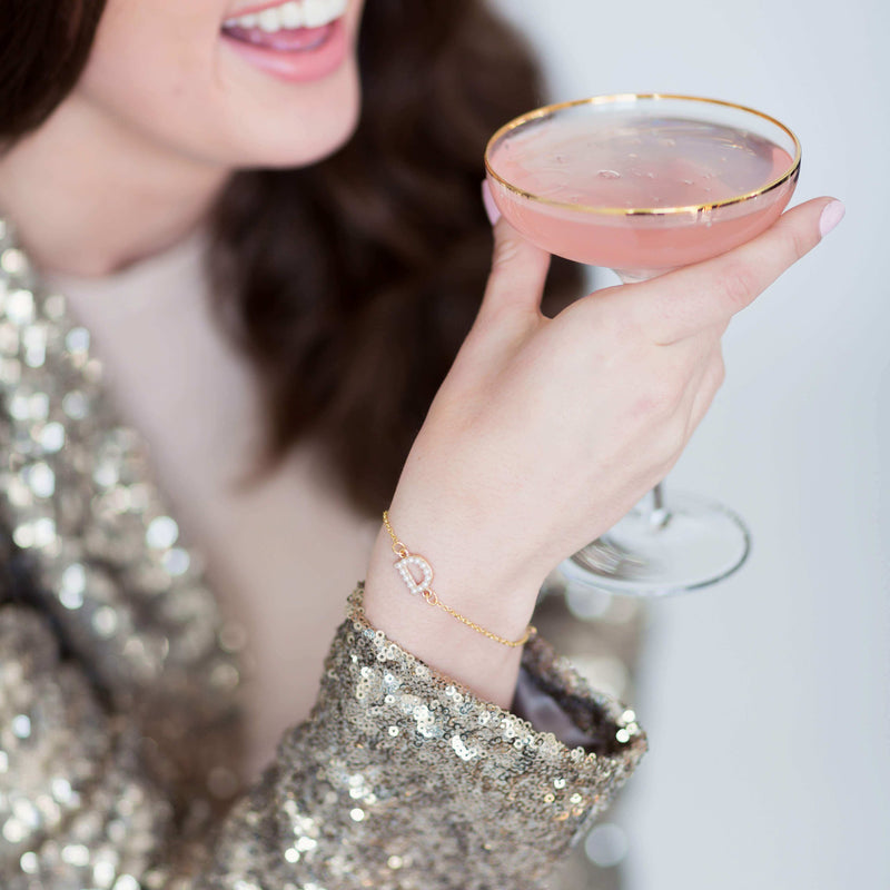 Models wears the personalised pearl initial bracelet on a gold chain with the pearl initial D while holding cocktail and laughing