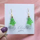 image shows green glass Christmas tree earrings with gold star detail on a white card that reads merry Christmas