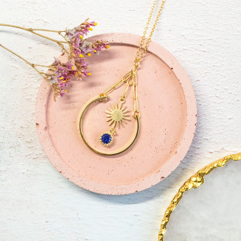 Image shows gold plated sunburst charm with a deep blue crystal attached, suspended within a golden arc. Necklace sits on a pink circular backdrop.