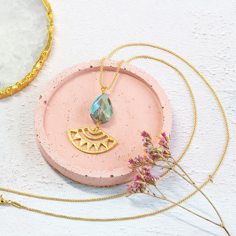 Image shows gold plated long length Andromeda necklace with a blue/green teardrop shaped crystal with a sun ray aztec charm. Necklace sits on a pink circular backdrop. 