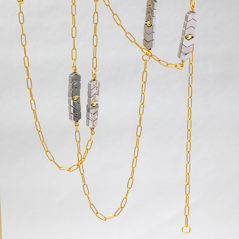 Image shows long length gold plated asteria necklace with four hematite chevron charms throughout. Necklace is spiralled on a white background.