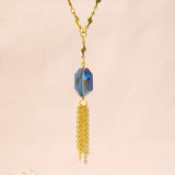 Image shows the blue iridescent hexagonal crystal with gold plated chain fringe suspended on a gold plated trace chain with tiny lightening bolt detail. Necklace is on a pink backdrop.