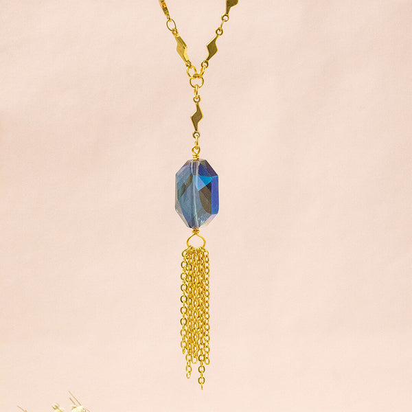 Image shows the blue iridescent hexagonal crystal with gold plated chain fringe suspended on a gold plated trace chain with tiny lightening bolt detail. Necklace is on a pink backdrop.