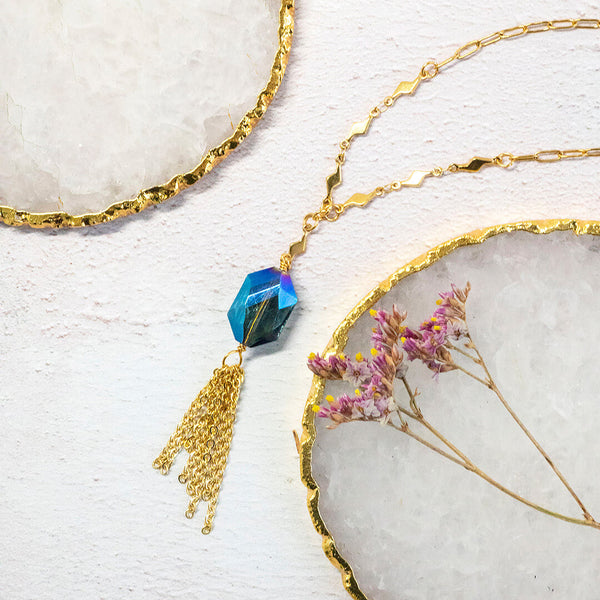 Image shows the blue iridescent hexagonal crystal with gold plated chain fringe suspended on a gold plated trace chain with tiny lightening bolt detail. Necklace is on a white backdrop.
