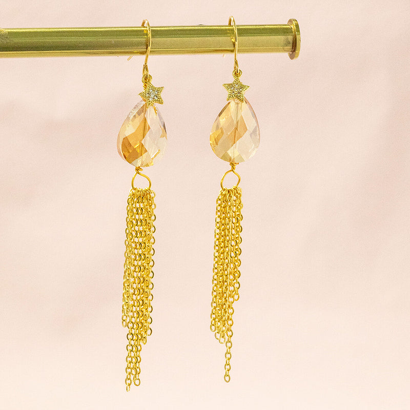 Image shows gold plated Canopus dangle earrings, complete with pave star and teardrop shaped topaz crystal with gorgeous golden tassels to complete the glam look. Earrings sit on a pink background.