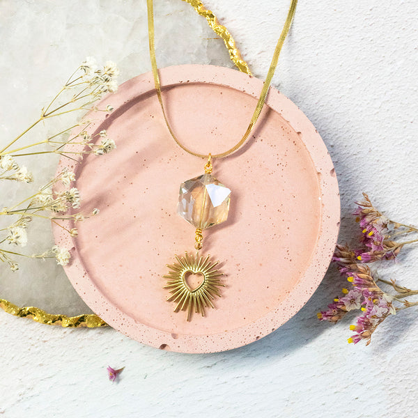 Image shows cosmic heart gold plated necklace with clear hexagonal crystal on a flat snake chain with heart sunburst charm. Necklace sits on a pink backdrop.