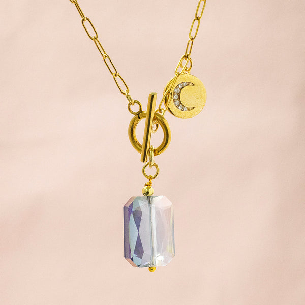 Image shows deimos gold plated necklace with front clasp and dainty crystal crescent moon disc charm. Necklace suspended in front of a pink backdrop.