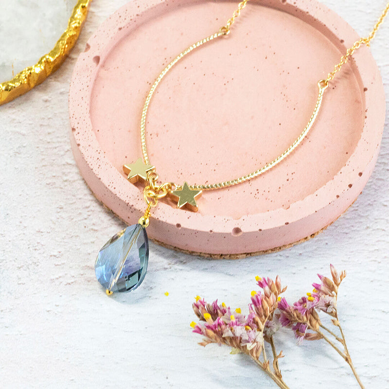 Image shows gold plated Elysium necklace on a pink and white backdrop. A gorgeous blue iridescent crystal suspended between two gold stars.