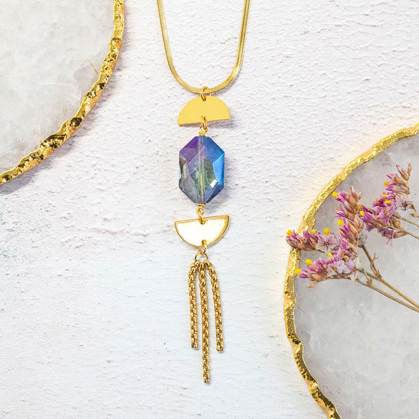 Image shows Epsilon gold plated necklace with gold chain tassels on a white backdrop. Crystal is a deep blue with an pink iridescence.