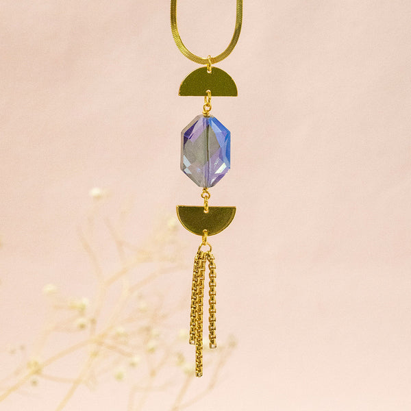 Image shows Epsilon gold plated necklace with gold chain tassel suspended in front of a pink backdrop. Crystal is a deep blue with an pink iridescence.