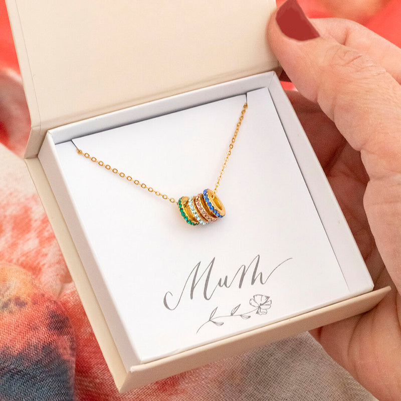 Image shows Gold plated family birthstone rings necklace on a 'Mum' sentiment card in the JOY branded gift box. Swarovski birthstone rings from left to right are May Emerald, March Aquamarine, June Light Amethyst and September Sapphire.