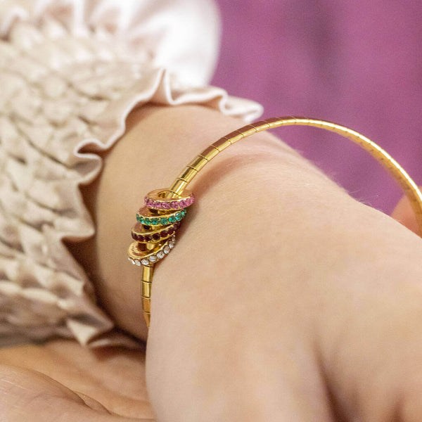 Model wears gold plated family birthstone rings bracelet with four different birthstone rings, from top to bottom as pictured: October Rose, May Emerald, July Ruby, April Crystal
