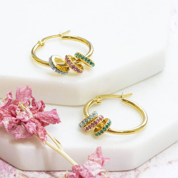 Image shows pair of gold plated birthstone hoop earrings on a white backdrop. Birthstone crystal rings from left to right: March Aquamarine, October Rose, May Emerald.   