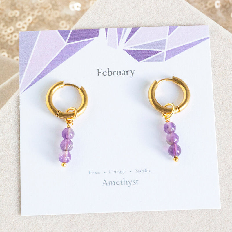 Image shows February amethyst earrings on a February sentiment card. Earrings are tiny gold plated huggie hoops with three tiny natural amethyst beads which can be removed.