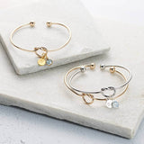 Image shows three Friendship Knot Bangles, one plain gold plated bangle with no charms, one silver plated bangle with initial disc charm with the letter 'H' engraved and a Swarovski birthstone charm in pale blue aquamarine March Birthstone and one gold plated with initial disc charm with the letter 'H' engraved and a Swarovski birthstone charm in pale blue aquamarine March Birthstone.
