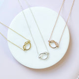Image shows flat lay of three Friendship Knot Necklaces, one gold plated, one silver plated and one rose gold plates. sat on a white circle and pink background.