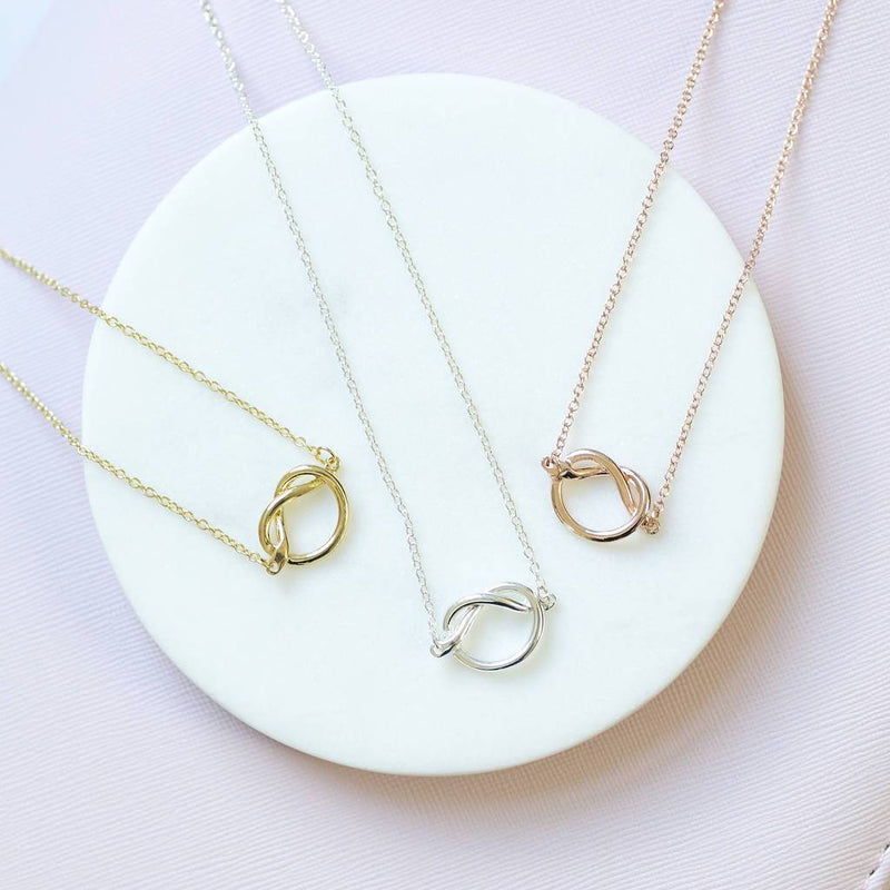 Image shows flat lay of three Friendship Knot Necklaces, one gold plated, one silver plated and one rose gold plates. sat on a white circle and pink background.