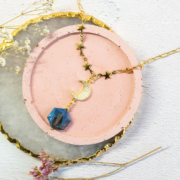 Image shows Galaxy Necklace with a gorgeous blue crystal, crystal encrusted crescent moon, and gold dainty star chain detail. Necklace sits on a pink backdrop. 