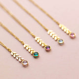 Image shows five gold leaf vine necklaces on a pink backdrop. Each with a different Swarovski birthstone. From left to right: April Crystal, May Emerald, February Amethyst, March Aquamarine, October Rose.