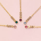Image shows five gold leaf vine necklaces on a pink backdrop arranged in a burst pattern. Each with a different Swarovski birthstone. From left to right: April Crystal, May Emerald, February Amethyst, March Aquamarine, October Rose.