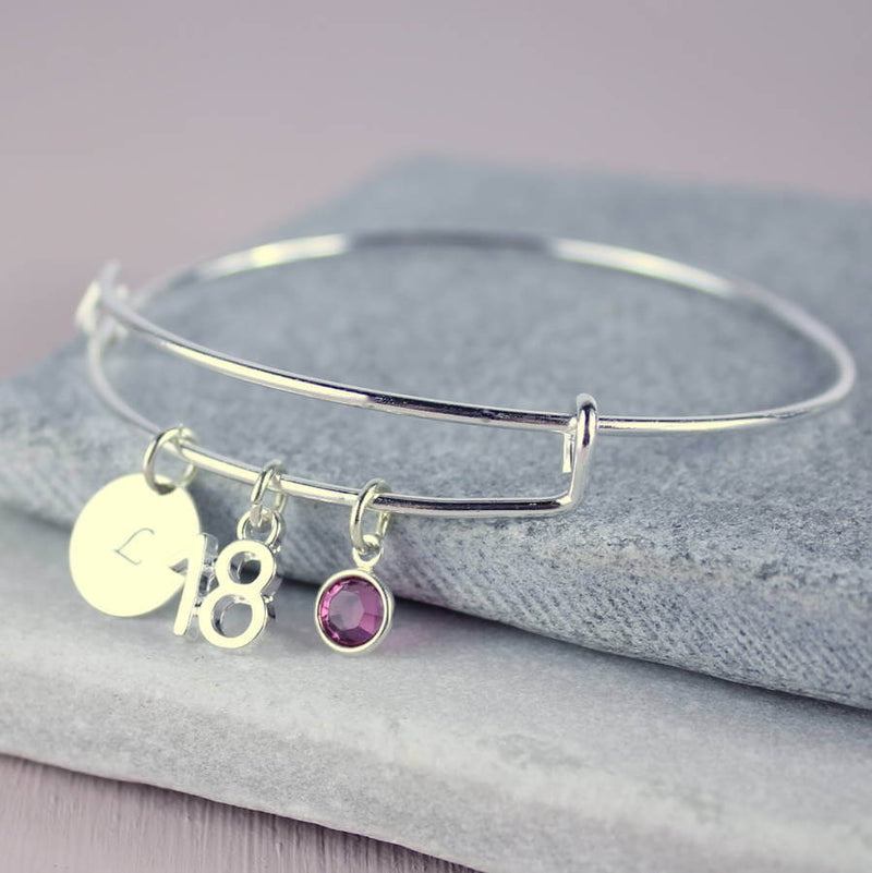 Image shows milestone birthday bangle with initial charm with the letter 'kL, '18' number charm and February Amethyst Swarovski crystal birthstone charm.