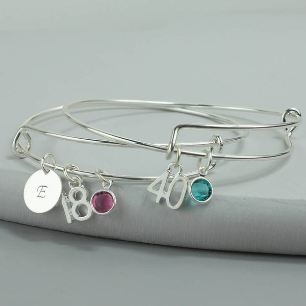 Image shows two milestone birthday bangles with initial charm with the letter 'E', '18' number charm and October Rose Swarovski crystal birthstone charm, second bracelet with '40' number charm and December Blue Zircon Swarovski crystal birthstone charm.