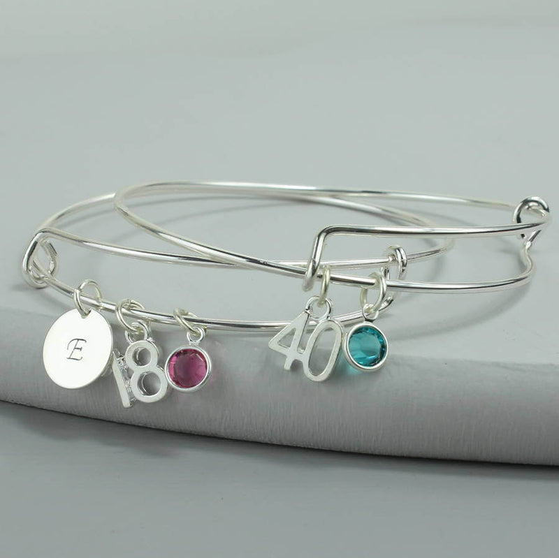 Image shows two milestone birthday bangles with initial charm with the letter 'E', '18' number charm and October Rose Swarovski crystal birthstone charm, second bracelet with '40' number charm and December Blue Zircon Swarovski crystal birthstone charm.