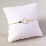 Image shows minimalist gold circle birthstone bracelet with May Emerald Swarovski Birthstone on a cream jewellery pillow.
