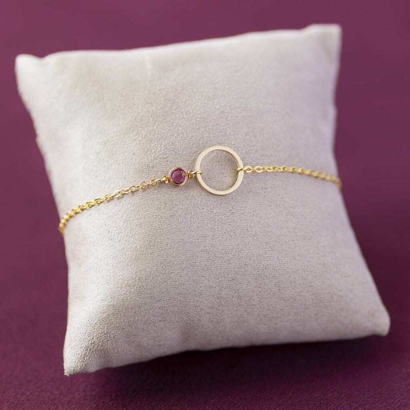 Image shows minimalist gold circle birthstone bracelet with October Rose Swarovski Birthstone on a cream jewellery pillow with a maroon backdrop.