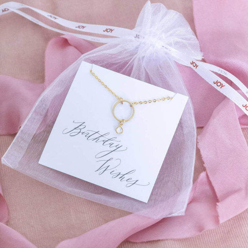 Image shows minimalist gold circle birthstone necklace with April Crystal Swarovski birthstone charm on a 'Birthday Wishes' sentiment card within the JOY organza gift pouch.