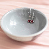 Image shows sterling silver mother and child birthstone earrings displayed in a grey ceramic bowl. Earrings show April crystal birthstone for child and January red garnet for mother.