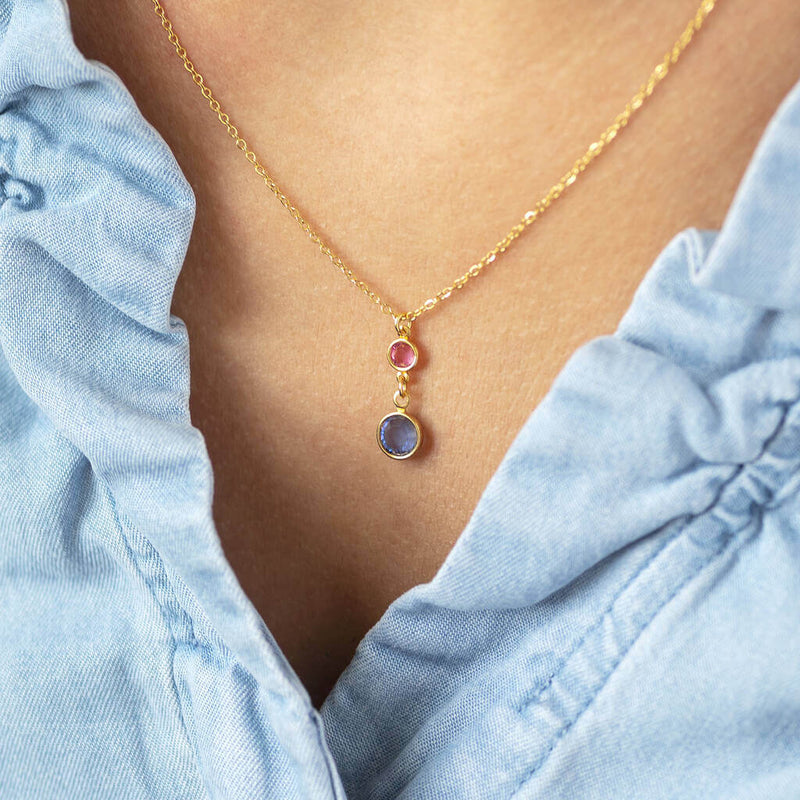 Model wears gold plated chain Mother and Child Birthstone necklace with two birthstones on one pendant. Top smaller birthstone to represent the child is October pink Rose. Larger birthstone to represent the mother is March aquamarine in pale blue. Model wears a denim blue top.
