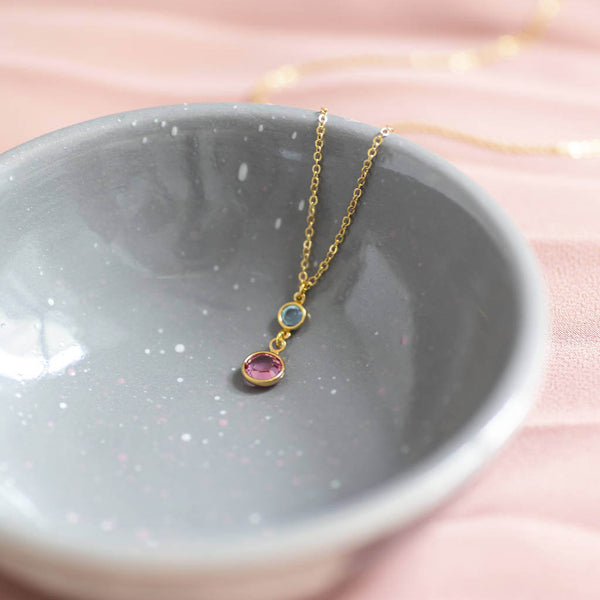 Image shows gold plated chain Mother and Child Birthstone necklace with two birthstones on one pendant. Top smaller birthstone to represent the child is October pink Rose. Larger birthstone to represent the mother is March aquamarine in pale blue. Displayed in a grey ceramic bowl in a pink backdrop.