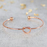Image shows Rose Gold Friendship Knot bangle with no birthstone or initial charms, on a grey backdrop.